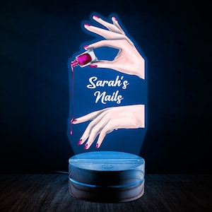 Puede incluir: Una luz nocturna impresa en 3D con un diseño de una mano sosteniendo una botella de esmalte de uñas con una gota de esmalte goteando sobre una uña. El texto "Sarah's Nails" está impreso en la luz.