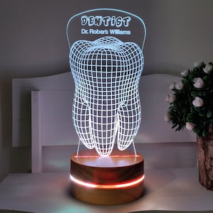 May include: A dentist-themed LED lamp featuring a 3D-rendered tooth design. The lamp has a wooden base with a glowing red and white accent. The top of the lamp reads "DENTIST" and "Dr. Robert Williams" in a stylized font. The lamp is a decorative item.