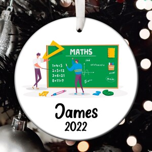 May include: A white round ornament with a green chalkboard featuring math equations and the word "MATHS" in black. Two cartoon figures are standing in front of the chalkboard. The ornament also includes the name "James" and the year "2022" in black.