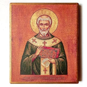 Op de afbeelding: Een rechthoekig houten icoon met een afbeelding van de heilige Augustinus van Hippo, bisschop en kerkleraar. De figuur heeft een gouden aureool, draagt groen-gouden gewaden met kruisen en houdt een rood boek vast. De tekst luidt SAINT AVGVSTINE OF HIPPO, BISHOP & DOCTOR.