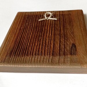 K&ouml;nnte beinhalten: Quadratische Holzplakette mit strukturierter, geriffelter Oberfl&auml;che. Das Holz hat eine warme braune Farbe mit einem helleren braunen Rand. Eine kleine Schnurschlaufe ist oben in der Mitte zum Aufh&auml;ngen befestigt. Die Plakette dient wahrscheinlich dekorativen Zwecken.