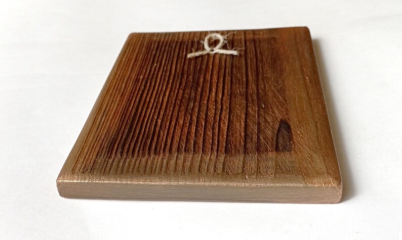 K&ouml;nnte beinhalten: Quadratische Holzplakette mit strukturierter, geriffelter Oberfl&auml;che. Das Holz hat eine warme braune Farbe mit einem helleren braunen Rand. Eine kleine Schnurschlaufe ist oben in der Mitte zum Aufh&auml;ngen befestigt. Die Plakette dient wahrscheinlich dekorativen Zwecken.