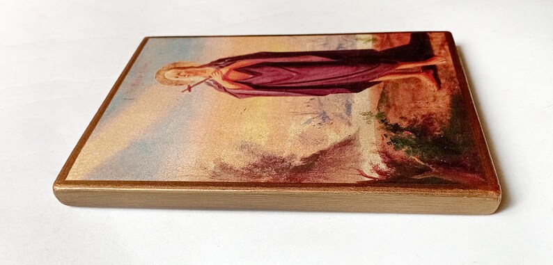 K&ouml;nnte beinhalten: Rechteckige Holzplakette mit religi&ouml;ser Malerei. Das Kunstwerk zeigt eine Figur in einem langen, weinroten Gewand, die ein Kreuz h&auml;lt. Der Hintergrund hat eine Landschaft mit warmen Erdt&ouml;nen und einem goldenen Rahmen.