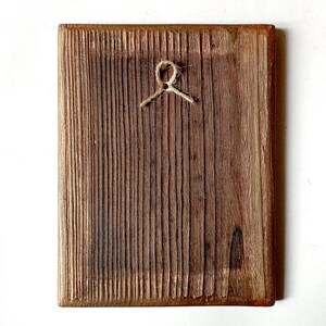 K&ouml;nnte beinhalten: Rechteckige Holzplakette mit strukturiertem, vertikalem Maserungsmuster. Oben ist eine nat&uuml;rliche Schnurschlaufe zum Aufh&auml;ngen angebracht. Das Holz hat einen warmen, braunen Farbton mit dunkleren Bereichen, was auf eine rustikale oder handwerkliche &Auml;sthetik hindeutet.