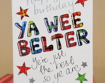 Op de afbeelding: Een witte verjaardagskaart met de tekst "happy birthday" in glinsterend blauw schrift. De woorden "YA WEE BELTER" staan in grote, kleurrijke geruite letters, met steraccenten.