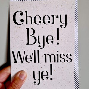 May include: A greeting card with the words "Cheery Bye! We'll miss ye!" in black font. The card has a speckled, off-white background and a striped border. The card is held by a hand.