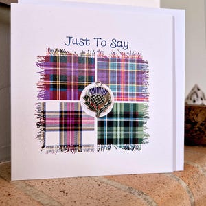 May include: A white card with the words "Just To Say" above four tartan squares. A thistle charm is attached. The plaid squares display purple, red, green, and white patterns, evoking Scottish heritage.