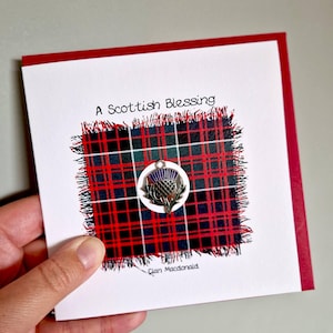 May include: A greeting card with a red border and a white front. The front features a red, black, and green tartan design with a silver thistle charm. The text "A Scottish Blessing" is printed at the top.