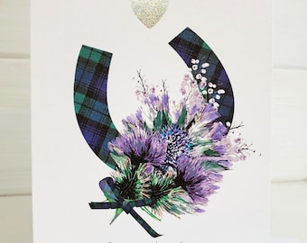 May include: A white wedding card with the text "On Your Wedding Day" and "So proud to be celebrating with you." The card features a silver glitter heart and a horseshoe shape with a tartan pattern. A bouquet of purple flowers is in the center.