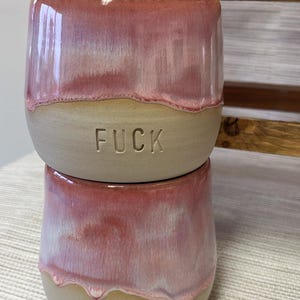 Könnte beinhalten: Zwei gestapelte Keramikbecher mit einer glatten, unglasierten beigen Basis und einer glänzenden, herablaufenden rosa Glasur im oberen Bereich. Jeder Becher ist auf der Vorderseite mittig im beigen Teil mit dem Wort FUCK in Großbuchstaben geprägt.