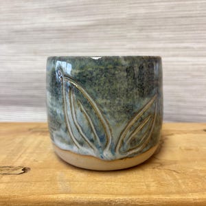 Könnte beinhalten: Eine handgefertigte Keramiktasse mit einem Blattdesign. Die Tasse ist zylindrisch mit einem hellbraunen Boden und einer dunkelblau-grünen glasierten Außenseite. Das Blattdesign ist in die Oberfläche geschnitzt und mit einer helleren Glasur hervorgehoben.