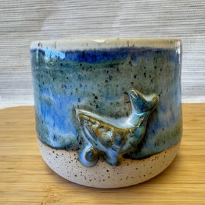 Könnte beinhalten: Ein Keramikbecher mit einem handgeformten Wal-Relief auf der Außenseite. Der obere Teil des Bechers ist in Blau- und Grüntönen mit dunklen Sprenkeln glasiert, während der untere Teil eine gesprenkelte cremefarbene Basis mit abgerundeter Form aufweist.