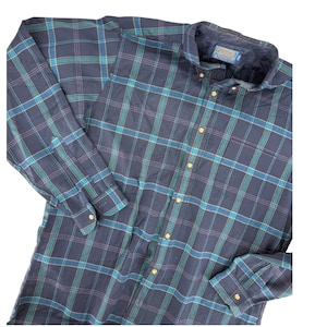 May include: A long-sleeved button-down shirt in a plaid pattern. The shirt features a navy blue base with teal, blue, and red lines. The shirt has a button-down collar and gold-colored buttons. The cuffs also have gold-colored buttons.