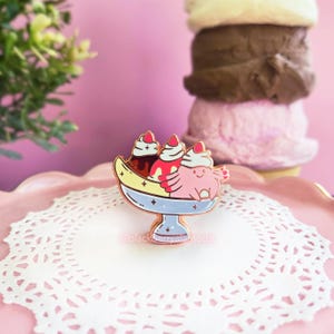 May include: A metal enamel pin shaped like a banana split dessert in a light blue dish. The pin features a pink axolotl, scoops of ice cream with whipped cream and cherries, and small star accents. The text @blackberryalamode is printed on the pin.