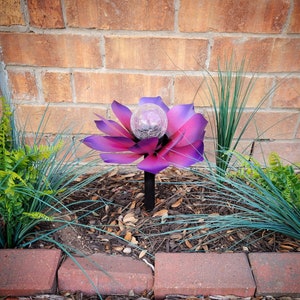 May include: A purple and pink metal flower with a clear glass globe in the center. The flower is on a black metal stake and is planted in a bed of brown mulch. The flower is surrounded by green grass and brick.