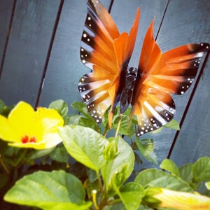 May include: A metal butterfly garden decoration with orange and black wings. The butterfly is perched on a green plant with yellow flowers.