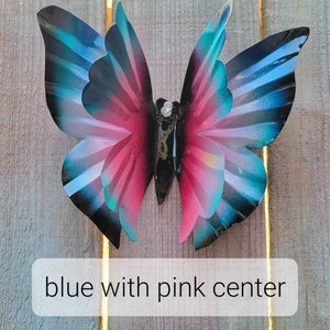 May include: A metal butterfly wall decoration with blue wings and a pink centre. The butterfly is attached to a wooden surface.