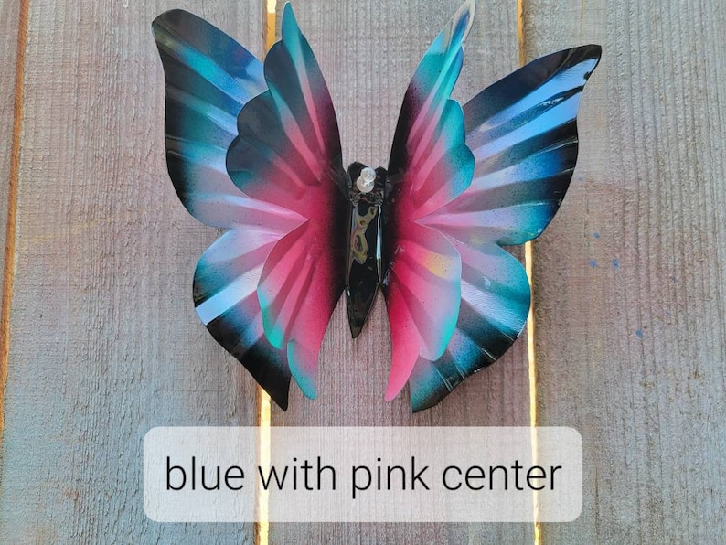 May include: A metal butterfly wall decoration with blue wings and a pink centre. The butterfly is attached to a wooden surface.