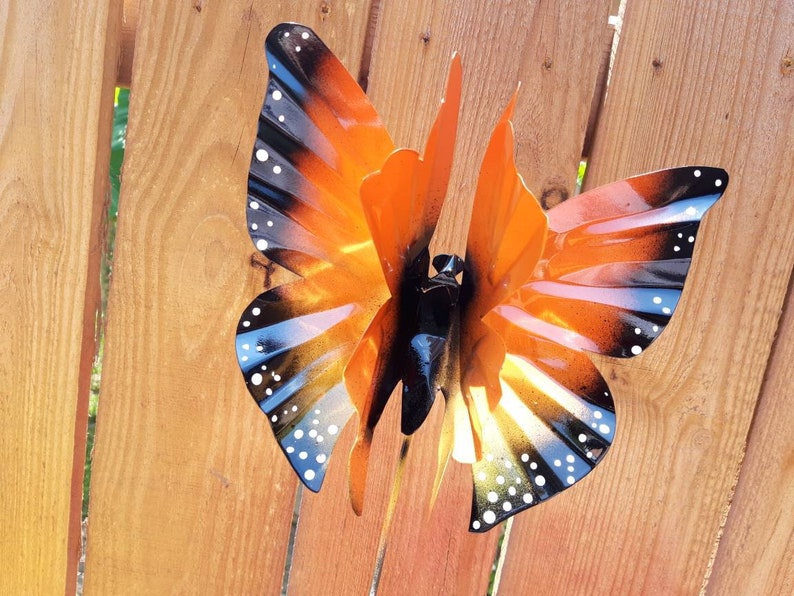 May include: A metal butterfly wind spinner with orange, black, and white paint. The butterfly has a detailed design with black and white spots on the wings.