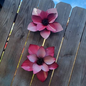 Puede incluir: Dos flores de metal rosa con un acabado texturizado est&aacute;n unidas a una valla de madera. Las flores tienen cinco p&eacute;talos cada una y un centro oscuro.