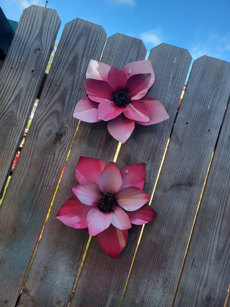 Puede incluir: Dos flores de metal rosa con un acabado texturizado est&aacute;n unidas a una valla de madera. Las flores tienen cinco p&eacute;talos cada una y un centro oscuro.