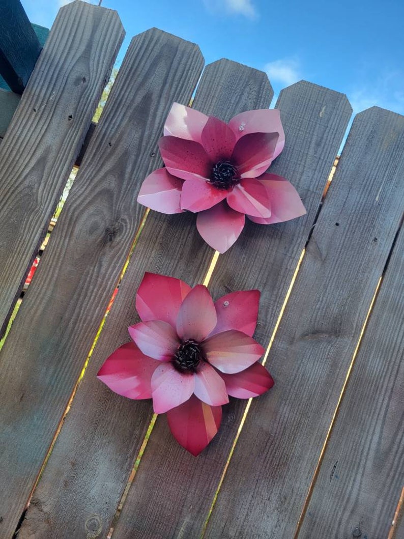 Puede incluir: Dos flores de metal rosa con un acabado texturizado est&aacute;n unidas a una valla de madera. Las flores tienen un dise&ntilde;o 3D y son de color rosa claro con un centro rosa m&aacute;s oscuro.