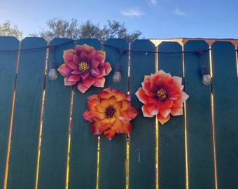 Puede incluir: Tres decoraciones de flores de metal en tonos rojos y naranjas cuelgan de una valla de madera verde. Las flores están unidas a una cadena de luces.