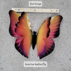 May include: A large metal sunrise butterfly with a black body and wings that are a gradient of orange, yellow, pink, and purple. The butterfly is on a grey concrete surface. "Size large" and "Sunrise butterfly" are written in black text on white rectangles below the butterfly.