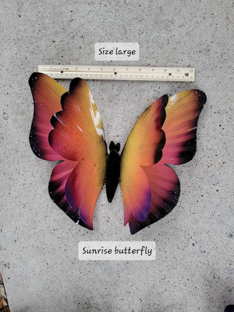 May include: A large metal sunrise butterfly with a black body and wings that are a gradient of orange, yellow, pink, and purple. The butterfly is on a grey concrete surface. "Size large" and "Sunrise butterfly" are written in black text on white rectangles below the butterfly.