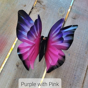 May include: A metal butterfly with purple and pink wings. The butterfly is painted with a gradient of colour, starting with a dark purple at the top and fading to a light pink at the bottom. The butterfly is attached to a wooden surface.