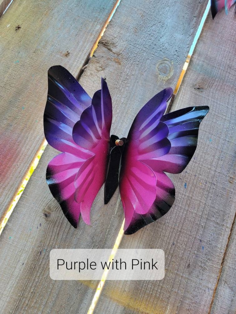 May include: A metal butterfly with purple and pink wings. The butterfly is painted with a gradient of colour, starting with a dark purple at the top and fading to a light pink at the bottom. The butterfly is attached to a wooden surface.