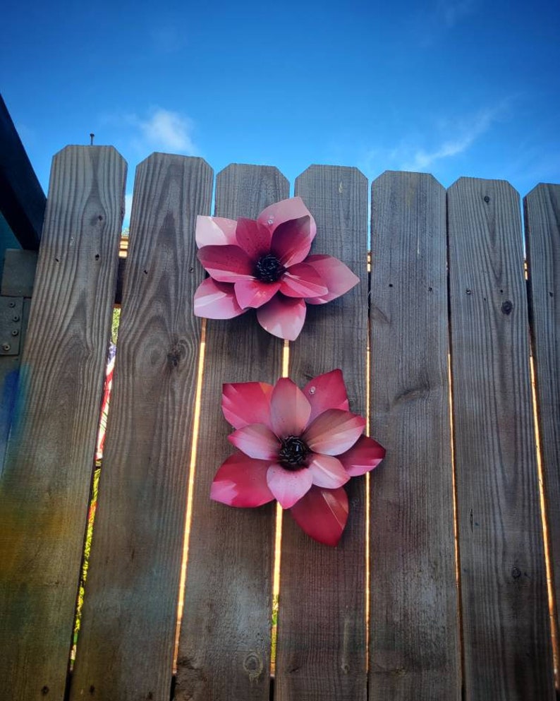 Puede incluir: Dos flores de metal rosa est&aacute;n unidas a una valla de madera. Las flores tienen un dise&ntilde;o 3D y est&aacute;n ligeramente elevadas de la superficie de la valla.