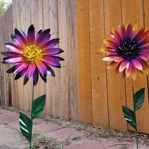 May include: Two metal flower garden stakes. One flower is purple, pink, and yellow, the other is orange, pink, and yellow. Both have green stems and leaves. The flowers are in front of a wooden fence.