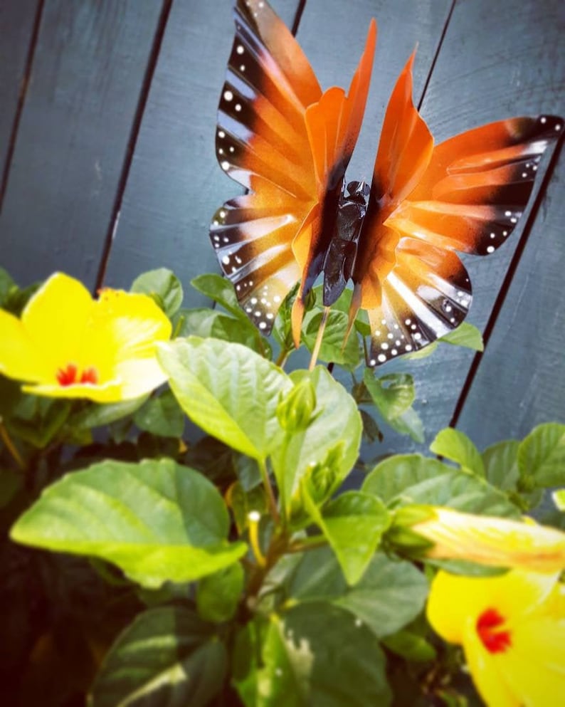 May include: A metal butterfly garden decoration with orange and black wings. The butterfly is perched on a green plant with yellow flowers.
