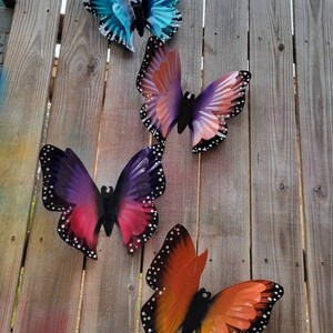 May include: Four metal butterflies in different colours and patterns are attached to a wooden fence. The butterflies are blue, purple, pink, and orange.