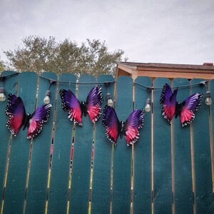 May include: A string of purple and pink butterfly decorations hanging on a green wooden fence with white string lights.