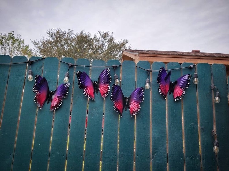 May include: A string of purple and pink butterfly decorations hanging on a green wooden fence with white string lights.