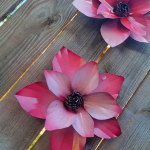 Puede incluir: Dos grandes flores de metal rosa con centros negros est&aacute;n unidas a una superficie de madera. Las flores tienen un degradado de color de un rosa m&aacute;s oscuro a un rosa m&aacute;s claro.