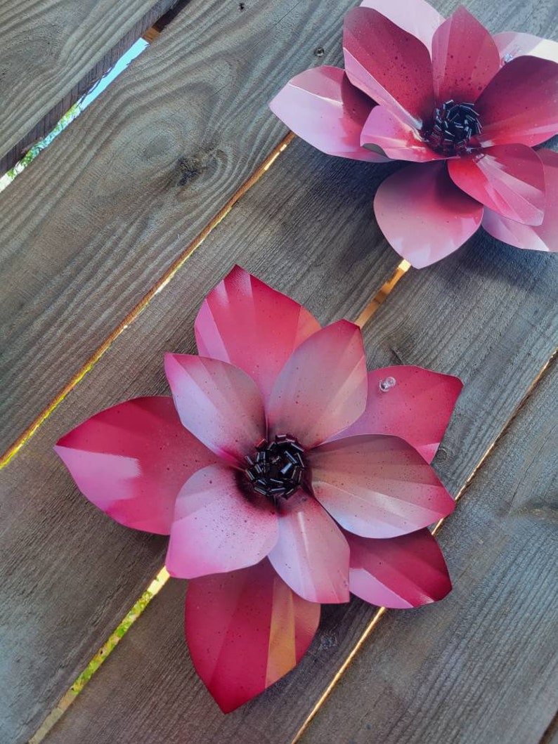 Puede incluir: Dos grandes flores de metal rosa con centros negros est&aacute;n unidas a una superficie de madera. Las flores tienen un degradado de color de un rosa m&aacute;s oscuro a un rosa m&aacute;s claro.