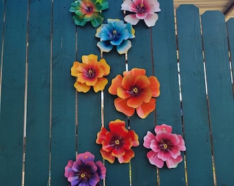 Puede incluir: Una valla de madera verde azulada con ocho decoraciones de flores de metal de colores. Las flores son en tonos de rojo, naranja, amarillo, azul, morado y verde.
