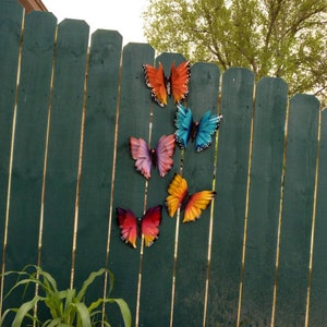 May include: Five colourful metal butterfly decorations are attached to a green wooden fence. The butterflies are in various shades of orange, yellow, blue, and purple.