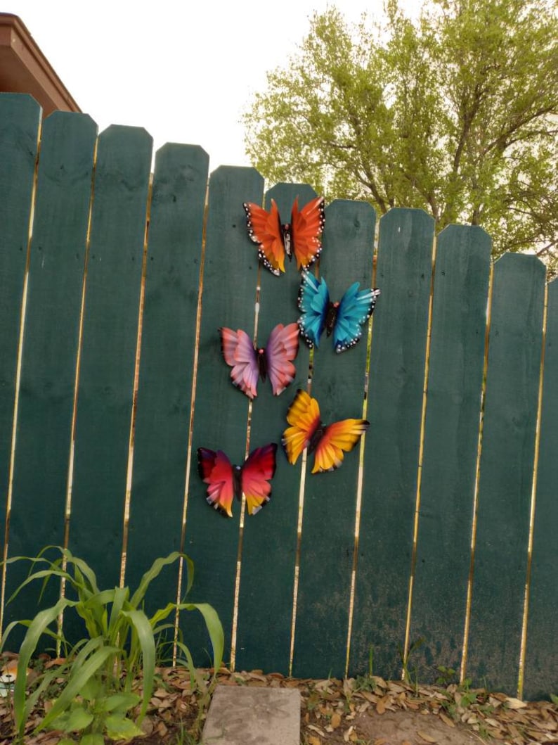 May include: Five colourful metal butterfly decorations are attached to a green wooden fence. The butterflies are in various shades of orange, yellow, blue, and purple.
