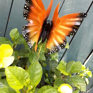 May include: A metal butterfly garden stake with orange and black wings and white spots. The butterfly is perched on a green plant with yellow flowers.