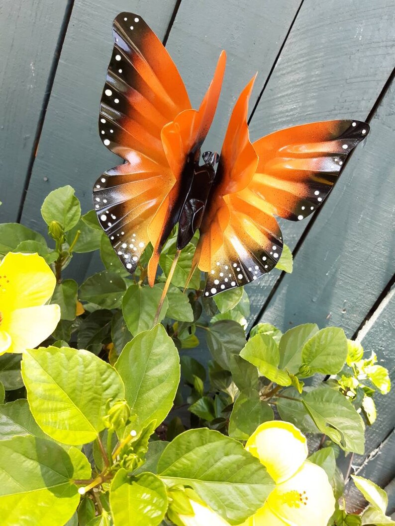 May include: A metal butterfly garden stake with orange and black wings and white spots. The butterfly is perched on a green plant with yellow flowers.