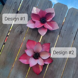 Puede incluir: Dos piezas de decoraci&oacute;n de pared de flores de metal en un esquema de color rosa y blanco. Las flores est&aacute;n unidas a una valla de madera. La flor superior est&aacute; etiquetada como "Dise&ntilde;o #1" y la flor inferior est&aacute; etiquetada como "Dise&ntilde;o #2".