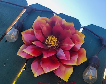 Puede incluir: Una gran escultura de flores de metal con pétalos rojos, rosas y amarillos. La flor está montada en una valla de madera verde, con luces de cadena y bombillas transparentes en la parte superior. El cielo es azul claro.