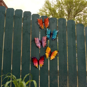 May include: A green wooden fence with five colourful metal butterflies attached to it. The butterflies are in various shades of blue, orange, yellow, and purple.