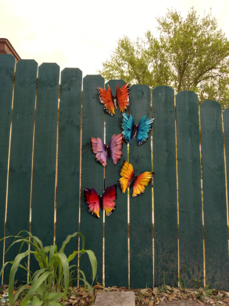 May include: A green wooden fence with five colourful metal butterflies attached to it. The butterflies are in various shades of blue, orange, yellow, and purple.