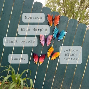 May include: Five metal butterfly wall decorations in various colours and designs. The butterflies are attached to a green wooden fence. The butterflies are labelled with their names: Monarch, Blue Morpho, light purple, Sunset, and yellow black centre.