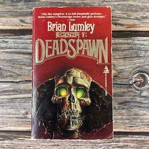 May include: A vintage paperback book titled "Necroscope V: Deadspawn" by Brian Lumley. The cover is predominantly red with gold lettering and features a detailed skull with glowing green eyes. The book is set against a rustic wooden background.